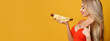Glamorous sexy blonde looks seductively at a hot dog holding in her hand. Woman posing on orange background with place for text. Commercial banner. Fast food advertising concept.