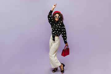 Wall Mural - Positive cool girl with curly hair, sunglasses and modern beret in polka dot long sleeve blouse and white trousers smiling.