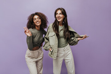 Joyful curly haired lady in modern to and light pants showing peace sign, smiling and posing with beautiful girl in olive jacket.