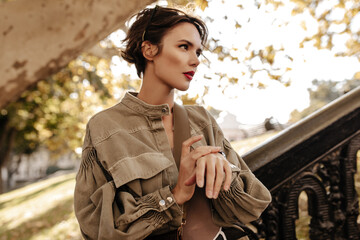 Wall Mural - Wonderful lady in wide denim jacket looking away outside. Trendy woman with short curly hairstyle and bright lips posing outdoors..