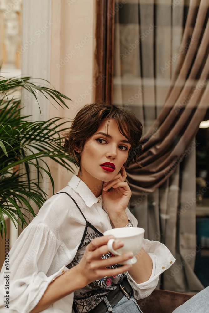Short-haired woman in long sleeve shirt with red lips holding cup of coffee in restaurant. Girl with brunette hairstyle poses in cafe.. - obrazy, fototapety, plakaty 