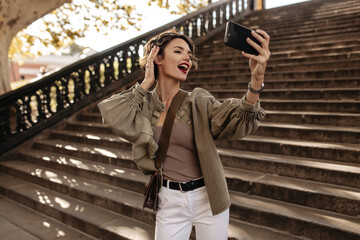 Wall Mural - Emotional girl in denim jacket and white jeans making selfie on background of stairs. Curly woman with handbag taking photo outside..