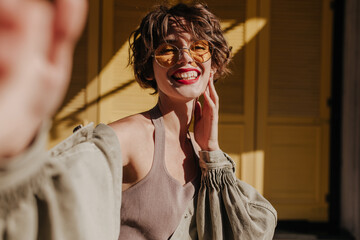 Wall Mural - Joyful woman in undershirt and olive jacket making selfie on bright backdrop. Short-haired girl with red lips smiles on yellow background..