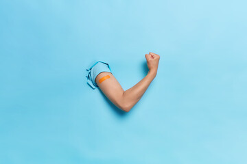 Horizontal shot of unrecognizable human keeps arms in paper hole clenches fist demonstrates power and strength after vaccination feels protected isolated over blue background shows bandage after shot