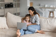 © fizkes - Smiling Latino mom and small teen daughter relax on couch at home use modern computer gadget together. Happy Hispanic mother and teenage girl child rest on sofa in living room have fun browse laptop.