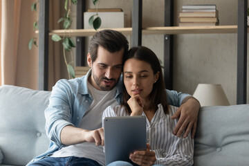 Wall Mural - Young couple sit on sofa spend carefree weekend time at home using tablet device. Family having fun using new mobile application, buying on-line, choose services goods on internet, make order concept