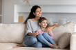 © fizkes - Happy Latino mother and biracial teen daughter sit relax on couch in living room cuddle and hug. Smiling young Hispanic mom and ethnic girl child rest on sofa at home look in distance dream embrace.