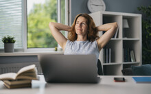 Pretty Female Freelancer Sitting On Comfy Chair With Hands Behind Head And Dreaming About Vacation. Mature Woman Using Laptop For Work A Home.