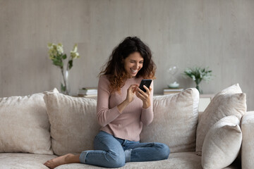 Poster - Overjoyed millennial Latino woman relax on sofa in living room talk speak on video call. Happy young Hispanic female rest on couch laugh have webcam virtual event on cellphone at home.