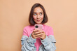 © WHstudio Leushin N - Dreamy young pretty Asian woman with dark hair holds mobile phone has thoughtful expression thinks about received message wears pink t shirt denim jacket looks away isolated over beige background.