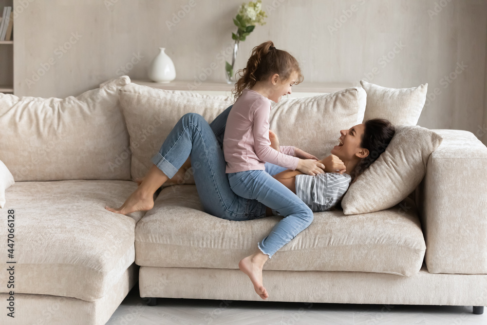 Small ethnic girl child and excited young Latino mom relax on couch in living room feel playful on weekend together. Overjoyed Hispanic mother and little daughter have fun play and tickle at home. - obrazy, fototapety, plakaty 
