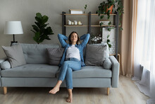 Serene Young Beautiful Woman Relaxing On Comfy Sofa With Hands Behind Head, Breath Fresh Conditioned Air Inside Of Modern Light Living Room. Modern House, Stress-free Weekend, Contemplation Concept