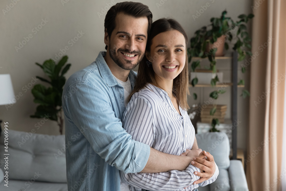Loving husband hugs beloved wife smiling look at camera enjoy harmonic relation, happy marriage, romantic relationships. Homeowners family, bank loan, medical insurance cover satisfied clients concept - obrazy, fototapety, plakaty 