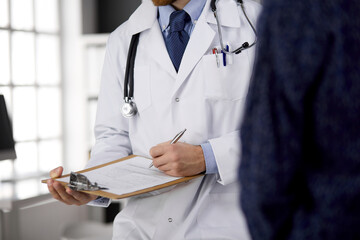 Unknown bearded doctor and patient woman discussing current health examination while sitting in sunny clinic, close-up. Medicine concept