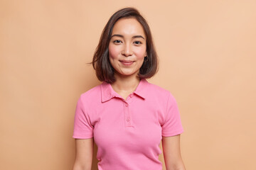 Wall Mural - Portrait of young Asian female model with natural beauty dark hair rouge cheeks healthy skin looks happily at camera wears casual pink t shirt isolated over beige background. People concept.