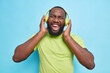 Joyful dark skinned man with thick beard keeps hands on headphones laughs happily enjoys favorite music wears casual green t shirt isolated over blue background. People and lifestyle concept