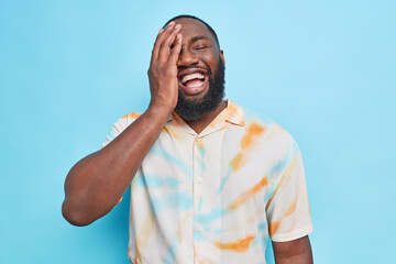 Wall Mural - Upbeat cheerful bearded Afro American man makes face palm laughs happily expresses happy authentic emotions dressed in washed out t shirt isolated over blue background laughs at something funny