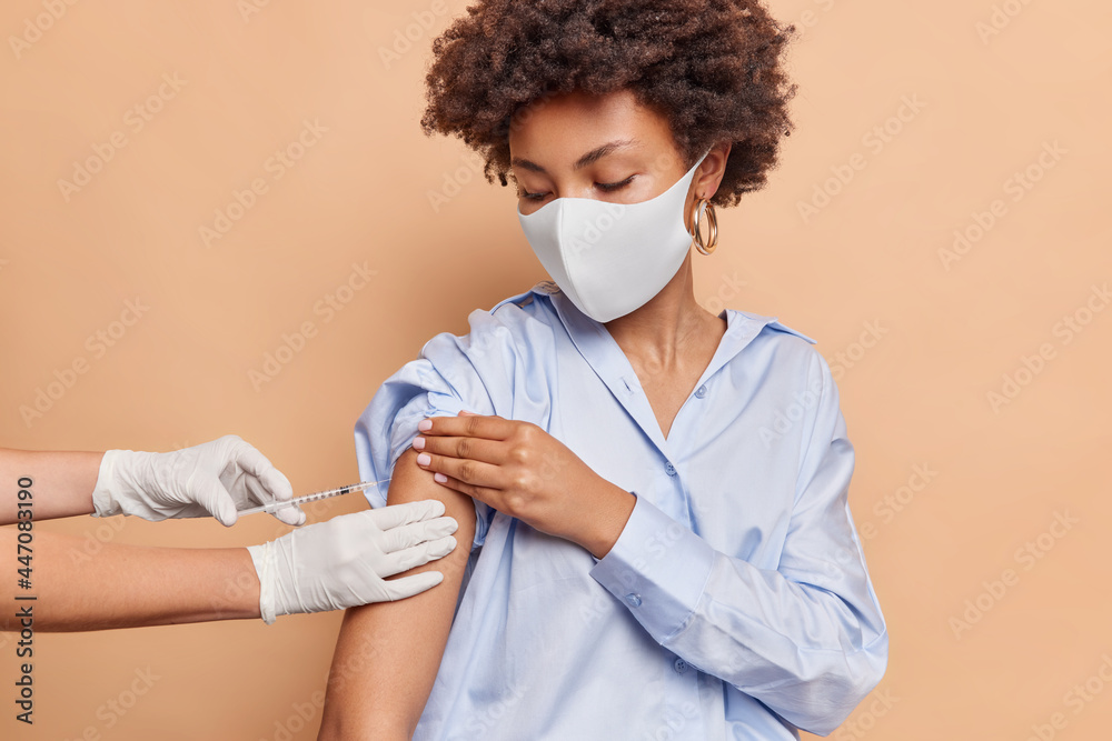 Coronavirus protection and mass immunization concept. Serious curly haired woman wears protective face mask against virus wears blue shirt receieves inoculation in shoulder isolated on beige wall - obrazy, fototapety, plakaty 