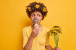 Unhappy woman has asthmatic attack problems with health wears oxygen mask which helps to breath holds wildflowers which cause allergic reaction has red swelling eyes isolated over yellow wall