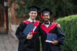 © Тарас Нагирняк - Portrait of two multinational graduate friends in graduation gowns with diploma in hands