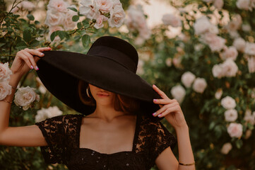 Elegant beautiful woman wearing, hiding her face under luxury wide brim black hat, posing near blooming roses. Copy, empty space for text
