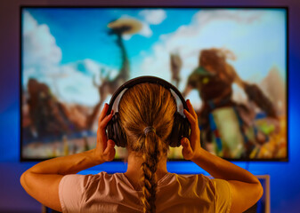 Wall Mural - Video games. Game strategy. Hobby. Gambling addiction. A young woman in headphones sits in front of a large computer monitor. She plays a video game. Close-up. Careful viewing. Shooting from the back.