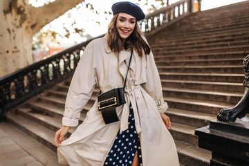 Wall Mural - Trendy young lady with brunette hair in beret, beige trench coat and crossbody bag, smiling and posing outdoors against old stairs background