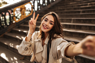 Wall Mural - Cute young brunette with red lips and beige trench coat sitting outdoors on old stairs, smiling and making selfie. Warm sunny autumn