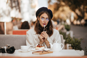 Wall Mural - Attractive young brown-haired girl in glasses, vintage beret and beige trench coat, chilling at city cafe terrace, eating cheesecake and tea, thinking about something