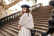 Photo from back of young brunette in dark beret and beige trench coat, posing on concrete stairs outdoors and looking into camera during daytime