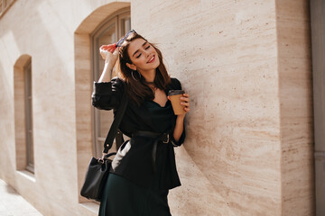 Wall Mural - Lifestyle portrait of charming young brunette with bright makeup, dark outfit with dress, jacket, bag, sunglasses and belt standing near beige wall outdoors and smiling