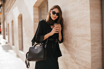 Wall Mural - Gorgeous young woman with brunette wavy hairstyle, sunglasses, black coat and bag walking in daylight city and posing against beige wall background