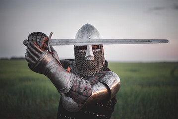 Antique knight in the plate armor and helmet holds a sword in hands close up.