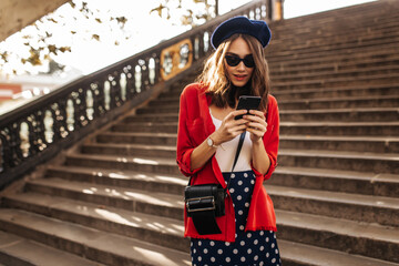 Wall Mural - Stylish young Parisienne with brunette wavy hair, beret, black sunglasses, white top, polka dot skirt and red shirt, searching something in phone outdoors. Old sunlit city stairs background