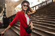 Charming young lady in trendy beret and sunglasses, with red shirt and black bag smiling and posing against warm autumn city background