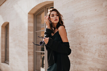 Wall Mural - Cute young girl with dark wavy hairstyle and bright makeup, silk dress, black jacket, holding sunglasses in hands and looking away against beige building background