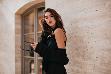 Wall Mural - Attractive young brown-haired lady with bright makeup, dark dress and black jacket posing outdoors against beige building background and looking into camera