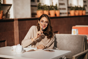 Wall Mural - Pretty young student girl with dark wavy hairstyle, trendy makeup, glasses and beige pullover, studying at city cafe terrace in autumn warm day. Notebook, phone, white teapot and cup on table