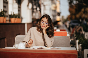 Wall Mural - Cute young student with brunette wavy hairstyle in beige sweater and glasses concentrated studying on city cafe terrace at sunny autumn day
