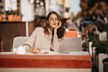 Wall Mural - Charming young brunette with red lips, glasses and beige sweater, learning something from notebook, having cup of tea at cafe terrace in warm sunny city