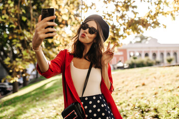 Wall Mural - Cute young girl with brown hair and red lips, in beret, black sunglasses, stylish top and shirt, making selfie outdoors. Sunny autumn park background