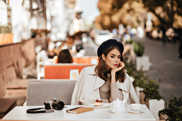 Wall Mural - Elegant young girl with brunette wavy hairstyle, vintage beret, beige trench coat having lunch at cafe terrace in warm sunny autumn city