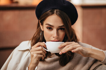 Wall Mural - Lifestyle portrait of elegant young woman with dark wavy hairstyle, trendy makeup, fashionable beige pullover and coat, sitting at cafe terrace and drinking coffee from white cup