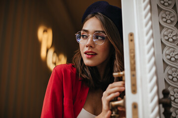 Wall Mural - Pretty young girl with brunette hair, stylish make-up, accessories, wearing beret, white and red clothes, holding door handle and looking away