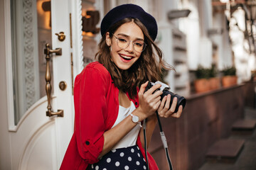 Wall Mural - Attractive pale girl with wavy dark hair, red lips, French style beret, red shirt and pretty accessories, smiling and making photo of autumn street