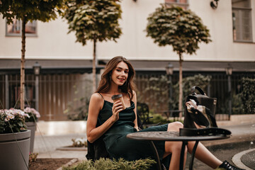 Wall Mural - Lifestyle portrait of charming young brunette in green silk dress sitting at city cafe terrace and drinking morning coffee. Light building and trees background