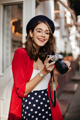 Wall Mural - Pretty young brunette with make-up, beret and glasses, wearing white top, red shirt and polka dot skirt, smiling and holding camera in hands at street