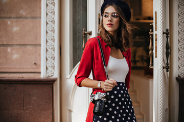 Wall Mural - Charming young photographer with brunette wavy hair in beret, white top, red shirt and polka dot skirt standing on street near cafe and looking away