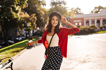 Wall Mural - Cute young brunette with beret, white top, red shirt and polka dot skirt, smiling, turning around and posing looking into camera against sunny city background