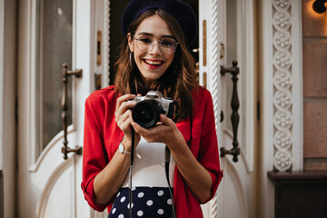 Wall Mural - Gorgeous young woman with red lips, glasses and beret on wavy dark hair, smiling, and making photo outdoors against background of light building